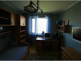 стул с плетеной спинкой у рабочего добротного стола с ящичками, навесная полка с глобусом, фотографиями и книгами на стене голубой спальни трехкомнатной квартиры в панельном доме
