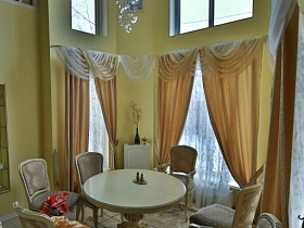 стулья со спинками вокруг белого круглого стола у эркерных окон с бежевыми шторами художественной дачи в сосновом лесу
