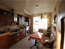 , коричневая мебельная стенка,обеденный стол с красной скатертью на резных ножках у дивана на светлой кухне с подвесным потолком и плиткой на полу простой просторной двухкомнатной квартиры с балконами