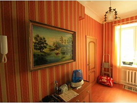 лестница-стремянка и красная спортивная сумка в углу у окна,большая картина в рамке и белый домофон на стене с полосатыми обоями над коричневым шкафчиком прихожей с люстрой на белом потолке