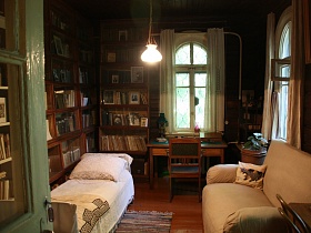 светлый диван с высокой спинкой, кровать с белым покрывалом, письменный стол у окна и шкафы с литературой в спальной комнате художественной деревянной дачи-музей времен СССР