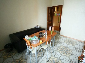 коричневый стол на белых ножках со стульями, черный прямоугольный диван и мультиварка рядом на полу со светло серой цветной плиткой кухни квартиры в переезде (въезде) молодоженов
