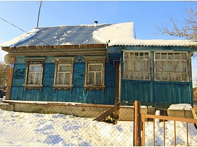 неокрашенные старые деревянные рамы на окнах голубого дома с верандой под шиферной крышей на участке под снегом в деревне