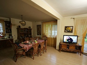 коричневая мебель кухни, обеденный стол со скатертью и стульями на светлом полу зонированной просторной комнаты семейного дома в глухом лесу