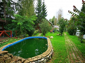бассейн с лестницей на лужайке с тропинками среди зеленых деревьев участка семейного дома в хвойном глухом лесу