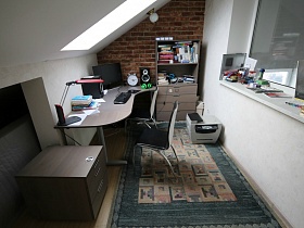 монитор, настольная лампа на поверхности белого стола, тумбочка, деревянная этажерка с книгами и бумагами, принтер на цветном коврике в рабочей комнате с окном на потолке